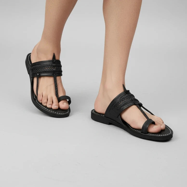 A pair of black leather thong sandals with braided straps and contrast white stitching, shown on a model's feet against a light gray studio background.
