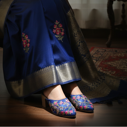 Woman in a blue saree with gold patterns sitting on a chair in a room.