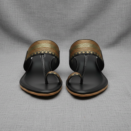 Black sandals with gold straps on a wooden surface surrounded by flowers and fabric.