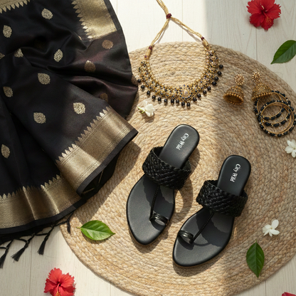 Black sandals with a black saree, necklace, and earrings on a woven mat with flowers.