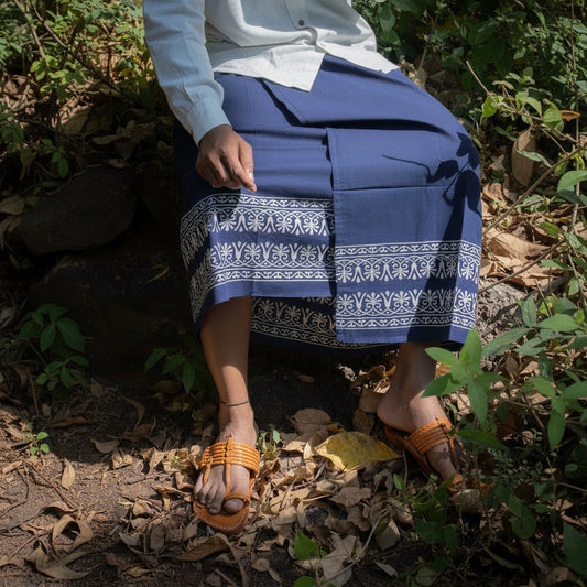 Person wearing a blue skirt with white patterns sitting on the ground among leaves and plants.