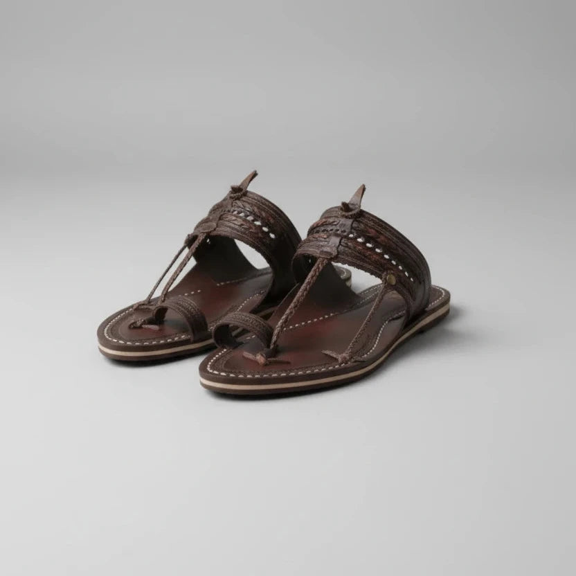 A pair of dark brown leather thong sandals with intricate braided straps and decorative stitching, resting on a wooden floor in warm natural light.