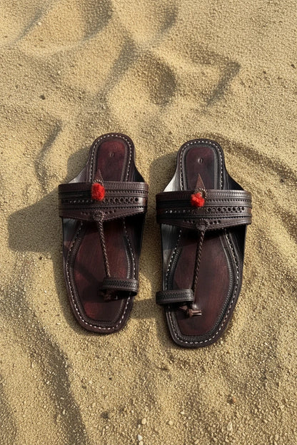 A pair of dark brown leather thong sandals with red pom-pom tassels, laid flat on sandy ground.