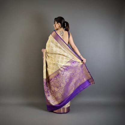 Woman holding a purple and gold saree in a room with a plant and chair.