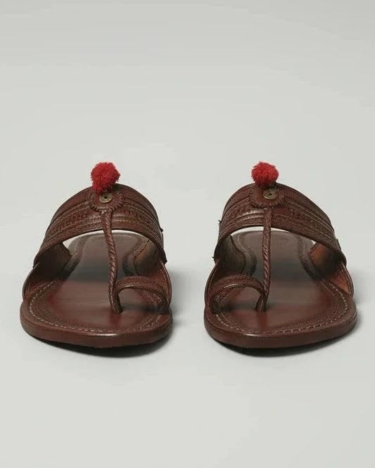 A pair of dark brown leather Thalori-style sandals with red tassels, shown on a person's feet wearing white pants on a beige tiled floor.