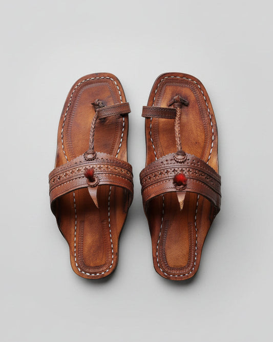 A pair of brown leather thong sandals with decorative stitched straps and small red bead details, laid flat on a textured gray concrete surface.