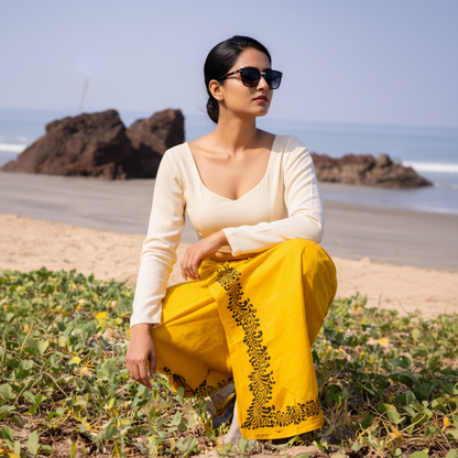 Woman in yellow traditional outfit with black patterns sitting on a beach.