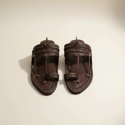 A pair of dark brown leather Moroccan-style sandals with intricate braided straps and a toe loop, displayed on a patterned rug.