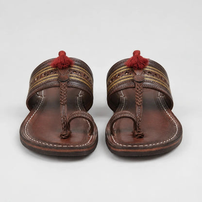A pair of brown leather thong sandals with decorative woven straps, white stitching, and red tassels on a light gray background.