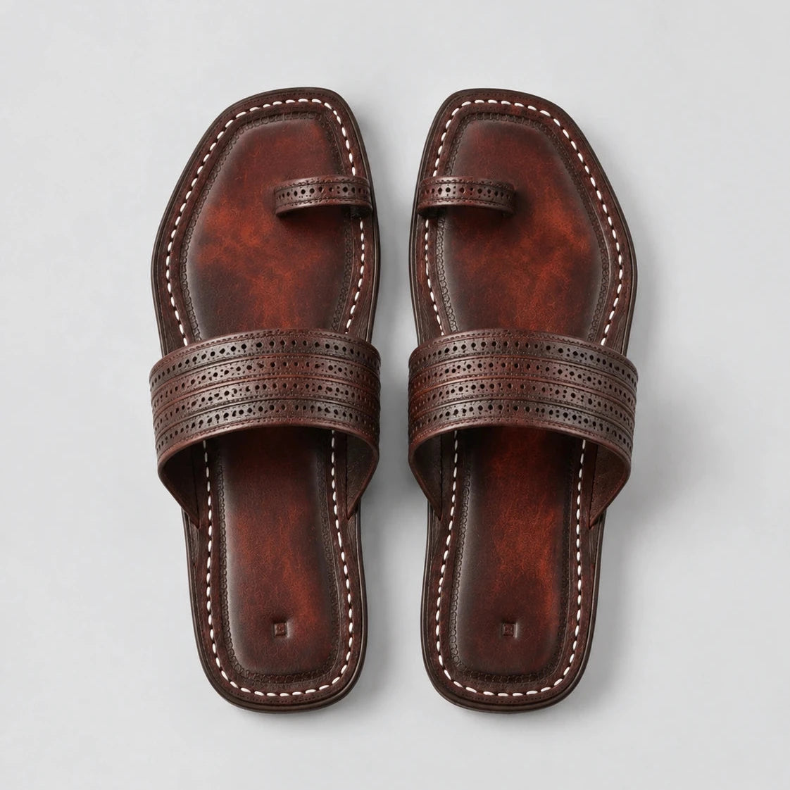 A pair of dark brown leather flat sandals with decorative perforated straps and contrast stitching, shown from a top-down view on a light gray background.